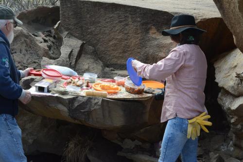Two people prepare food on a rocky surface, with various dishes and ingredients laid out around them.