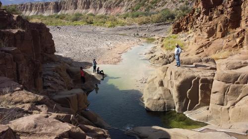 A group of people near a shallow stream surrounded by rocky terrain and dry landscape under a clear sky.