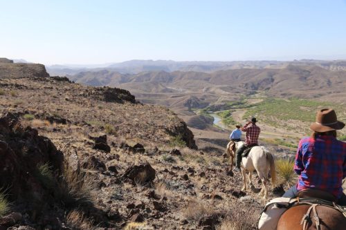 Two horseback riders navigate a rocky terrain with a vast, arid landscape and winding river in the background.
