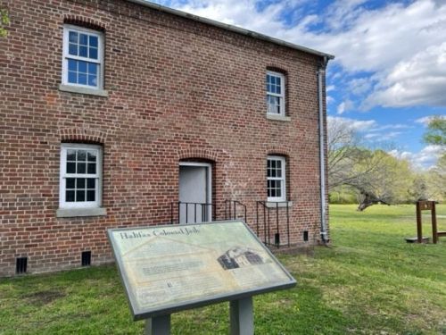 Historic brick building with windows, featuring a sign about Halifax Colonial Jail in a grassy area under a blue sky.