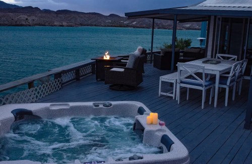 A serene lakeside deck features a hot tub, candles, and a cozy seating area with a fire pit, under a twilight sky.