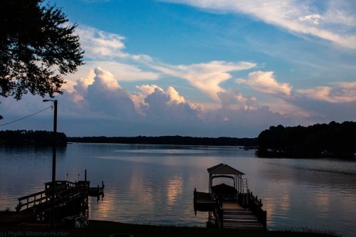 A serene lake at sunset, with fluffy clouds reflecting in the water and a dock extending into the scene.