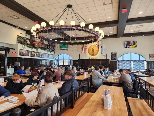 A bustling restaurant interior with wooden tables, patrons dining, and a large chandelier overhead.