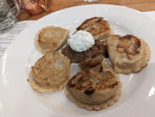 A plate of six golden-brown dumplings topped with sour cream and served with sautéed onions.