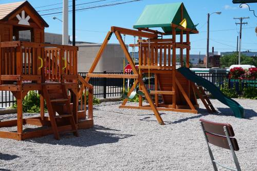 A wooden playground with a slide, swings, and a playhouse, set on a gravel surface in a sunny outdoor area.