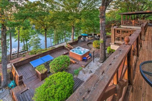A scenic outdoor space featuring a hot tub, ping pong table, and lush greenery by a lake, with wooden decks and seating areas.
