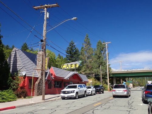 A red cafe building with a sloped roof, surrounded by trees, alongside a street with parked cars and an overpass above.