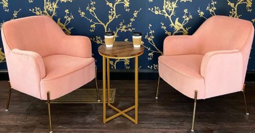 Two pink chairs facing each other with a small wooden table in between, set against a blue wall with gold floral patterns.