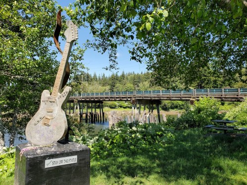 A guitar sculpture stands in a lush green area, with a bridge and water in the background.