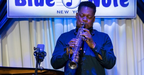A musician plays the saxophone on stage at Blue Note, with blue lighting and a piano in the background.