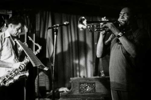 A black and white photo of two musicians performing on stage, one playing trumpet and the other on saxophone.