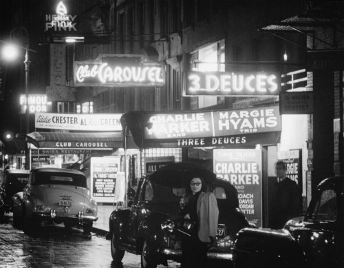 A vintage black-and-white street scene featuring neon signs for Club Carousel and 3 Deuces, with classic cars parked nearby.