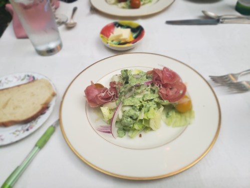A plate of salad with greens, cherry tomatoes, prosciutto, and a creamy dressing, accompanied by a glass of water and bread.
