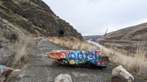 A graffiti-covered rock on a rocky path beside a river, surrounded by hills and a cloudy sky.