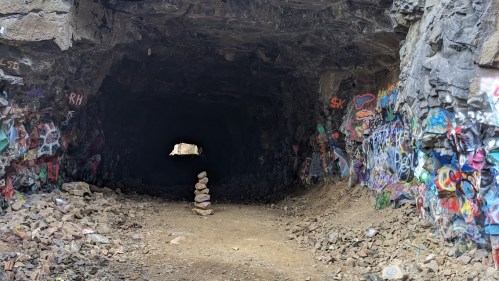 A dark tunnel with colorful graffiti on the walls and a stack of stones in the center, leading to a bright opening.