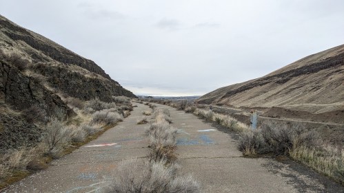 A deserted, cracked road surrounded by dry hills and sparse vegetation under a cloudy sky.