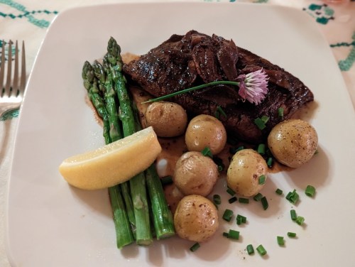 A plate of steak with asparagus, baby potatoes, a lemon wedge, and a flower garnish on a white background.