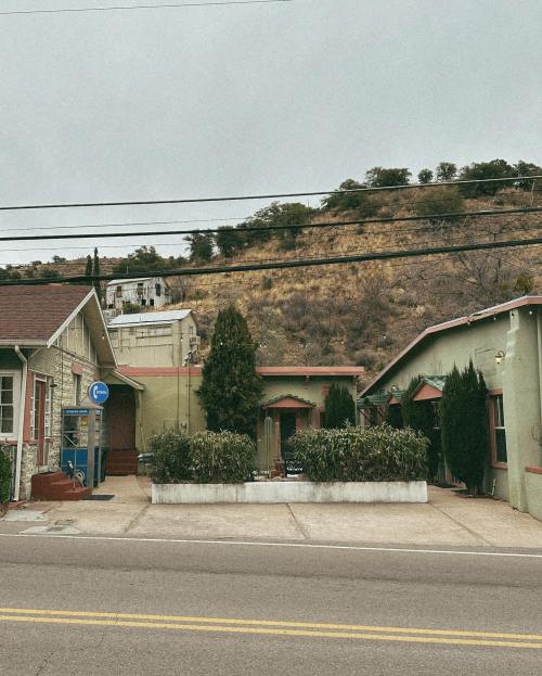 A street view of two green buildings with shrubs, set against a hillside under a cloudy sky. Power lines are visible.