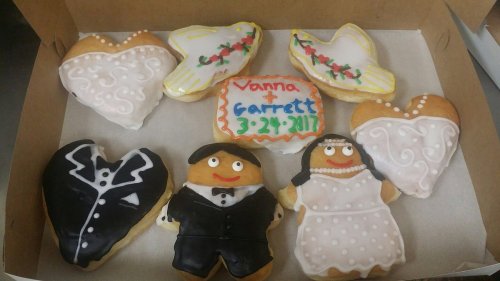 A box of decorated cookies featuring wedding-themed designs, including hearts, a bride and groom, and a date.
