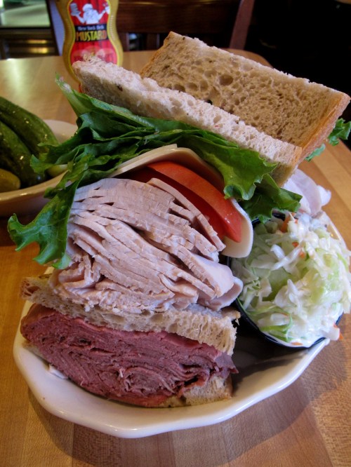A towering sandwich with layers of turkey, roast beef, lettuce, tomato, and coleslaw, served with pickles on the side.