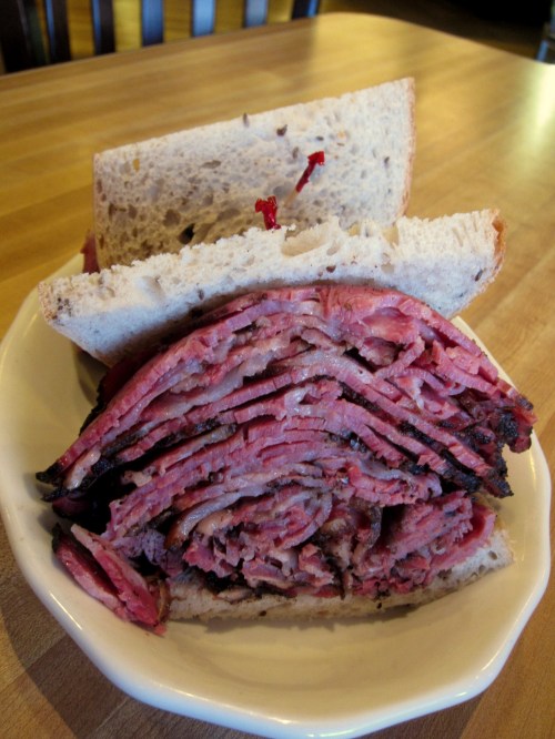 A close-up of a stacked pastrami sandwich on rye bread, served on a plate with a wooden table background.