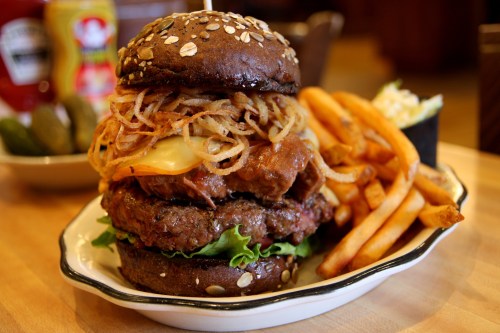A towering burger with two patties, cheese, crispy onions, lettuce, and a dark sesame seed bun, served with fries.