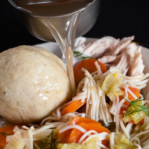 A bowl of chicken soup with noodles, vegetables, and a large dumpling, with broth being poured from a ladle.