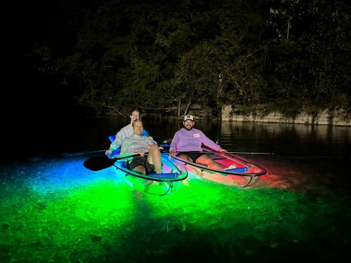 Three people in kayaks illuminated by colorful lights, paddling on a calm, dark waterway surrounded by trees.