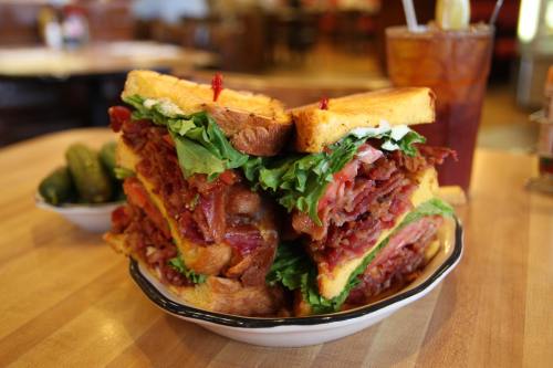A towering sandwich with layers of bacon, lettuce, and tomato, served with pickles and a drink in the background.
