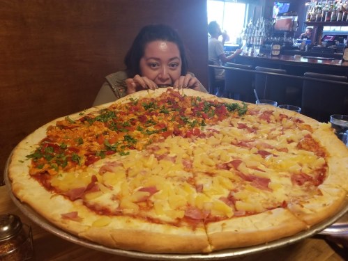 A person peeks over a giant pizza divided into two halves, one with toppings and the other with pineapple and ham.