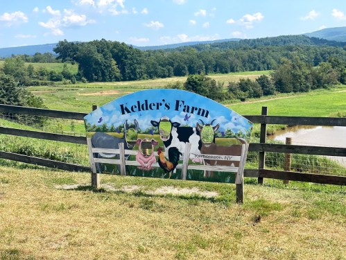 A colorful sign for Kelder's Farm featuring cartoon animals, set against a scenic rural landscape.