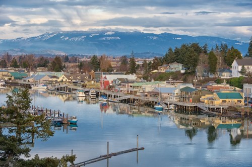 A serene waterfront scene with colorful houses, boats, and mountains in the background, reflecting in calm water.