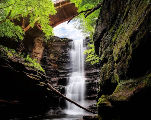 A serene waterfall cascades down rocky cliffs, framed by lush greenery and a bridge overhead.