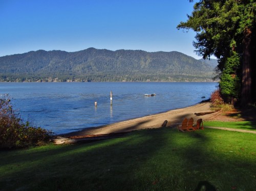 A serene lakeside view with mountains in the background, sandy shore, and green grass under a clear blue sky.