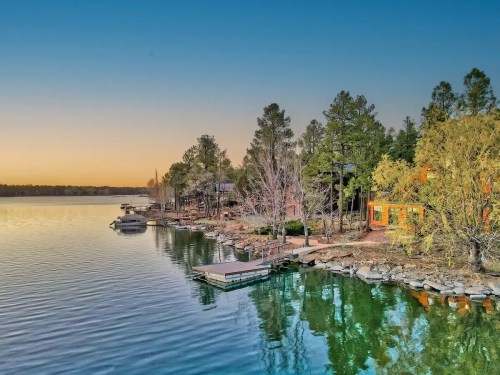 A serene lakeside view at sunset, featuring docks, boats, and trees reflecting on calm water.