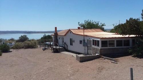 A white house with a metal roof near a lake, surrounded by sparse vegetation and clear blue skies.