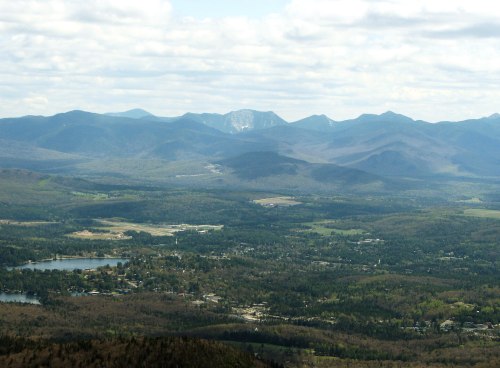 A panoramic view of mountains and valleys under a partly cloudy sky, with a lake and small town visible below.