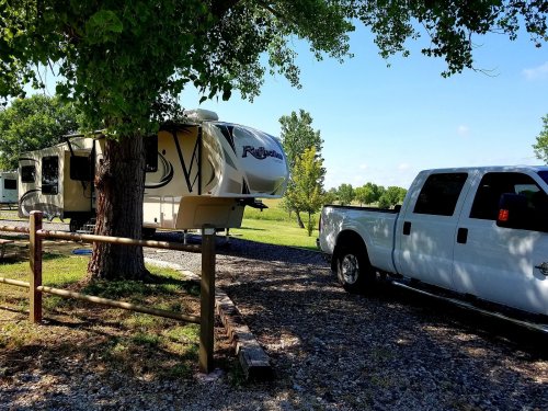 A white truck parked next to a large RV under a tree, with a clear blue sky and green landscape in the background.