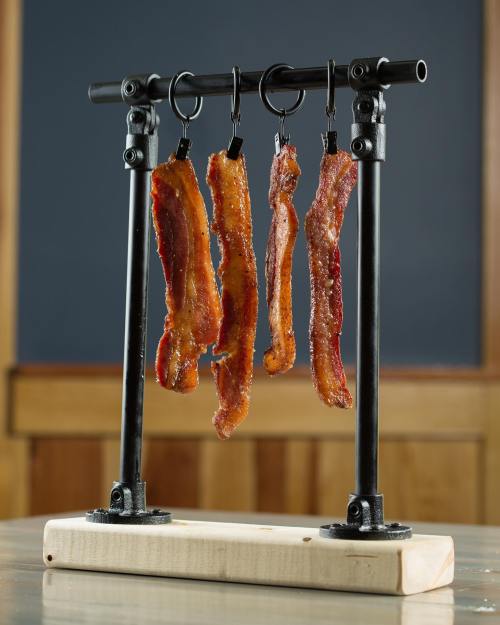 Four strips of crispy bacon hanging from a metal rack on a wooden base, set against a dark background.