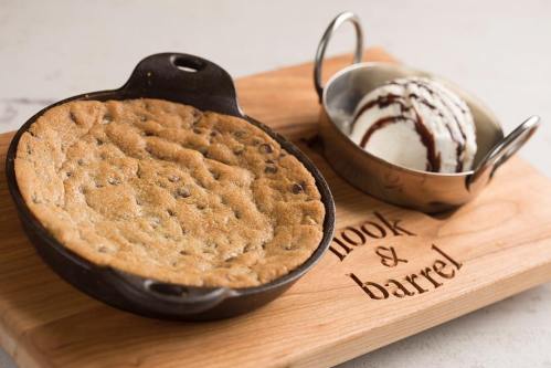 A warm chocolate chip cookie in a skillet, served with a scoop of ice cream and chocolate sauce on a wooden board.