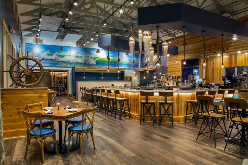 Modern restaurant interior featuring a bar, wooden decor, and nautical elements, with tables and chairs arranged for dining.
