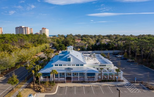 Aerial view of a blue-roofed building surrounded by trees and a parking lot, with distant buildings and ocean visible.