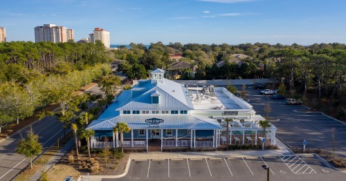 Aerial view of a blue-roofed restaurant surrounded by trees and a parking lot, with buildings in the background.