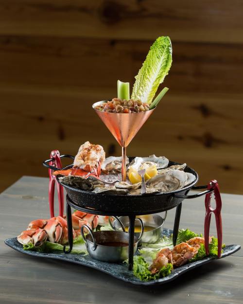 A seafood platter featuring oysters, shrimp, and a cocktail glass with garnishes, served on a decorative stand.