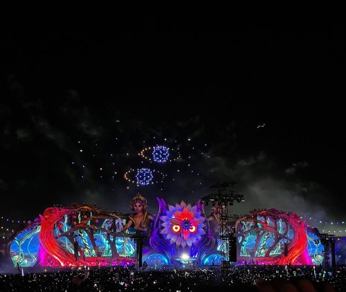 A vibrant festival stage illuminated at night, featuring colorful designs and fireworks in the sky.