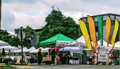 A vibrant farmers market with colorful flags, tents, and a "Road Closed" sign on a cloudy day.