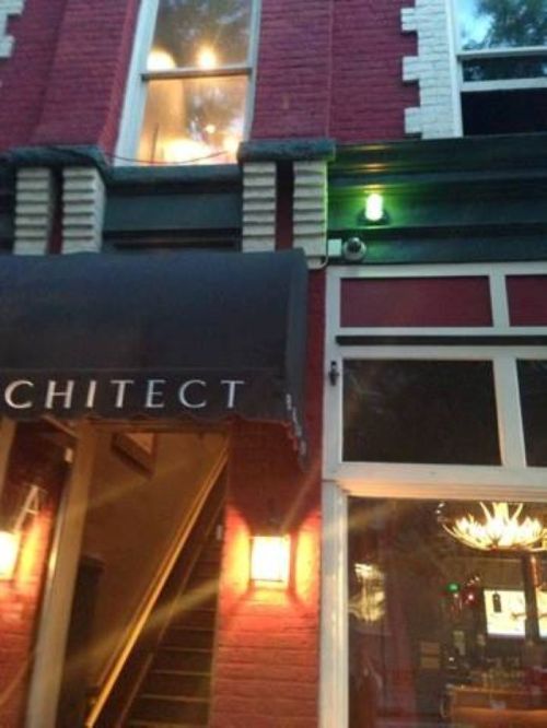 A red brick building with a black awning labeled "ARCHITECT" and warm lights illuminating the entrance.