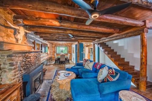 Cozy log cabin interior featuring a blue sofa, stone fireplace, wooden beams, and a staircase. Warm and inviting atmosphere.
