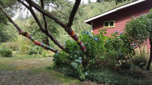 A colorful yarn-wrapped tree trunk beside a red house and blooming hydrangeas in a lush garden setting.