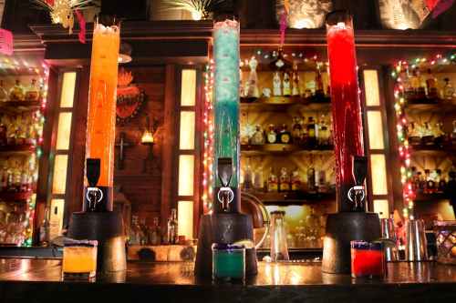 Three colorful drink dispensers filled with vibrant cocktails, set against a bar with shelves of liquor and festive lights.